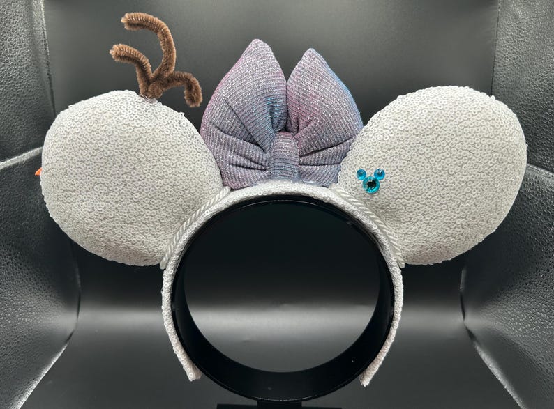 May include: A festive headband featuring white sequined ears, a shimmering bow with blue and purple hues, and a brown pipe cleaner antler. A blue jewel is on one ear. The headband is white with a black interior.