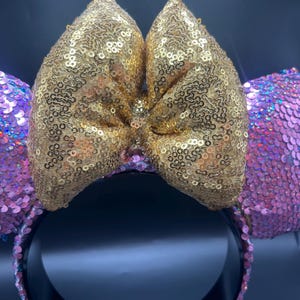 May include: A headband featuring two circular ears covered in iridescent pink sequins and a large central bow covered in shimmering gold sequins. The headband base is black, providing a contrast to the bright, reflective sequined fabric.