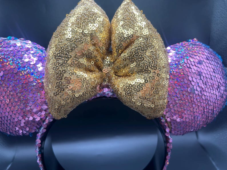 May include: A headband featuring two circular ears covered in iridescent pink sequins and a large central bow covered in shimmering gold sequins. The headband base is black, providing a contrast to the bright, reflective sequined fabric.