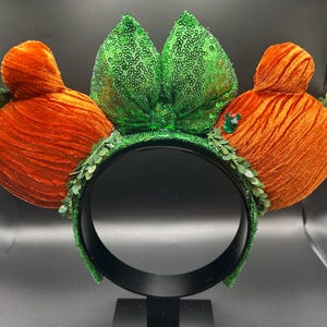 May include: A whimsical headband featuring orange velvet carrot-shaped ears and a central green sequined bow. The headband is adorned with green leaf accents and a black band, perfect for themed events or costumes.