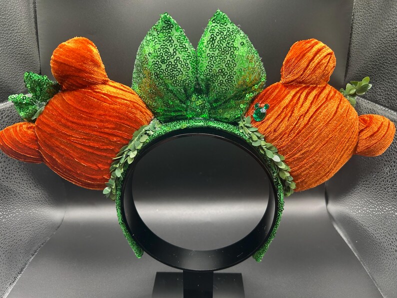 May include: A whimsical headband featuring orange velvet carrot-shaped ears and a central green sequined bow. The headband is adorned with green leaf accents and a black band, perfect for themed events or costumes.
