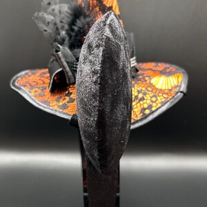 May include: A black velvet headband with a small witch hat design. The hat features orange and black lace detailing, a black feather, and a pointed top. The accessory is likely for Halloween or costume events.
