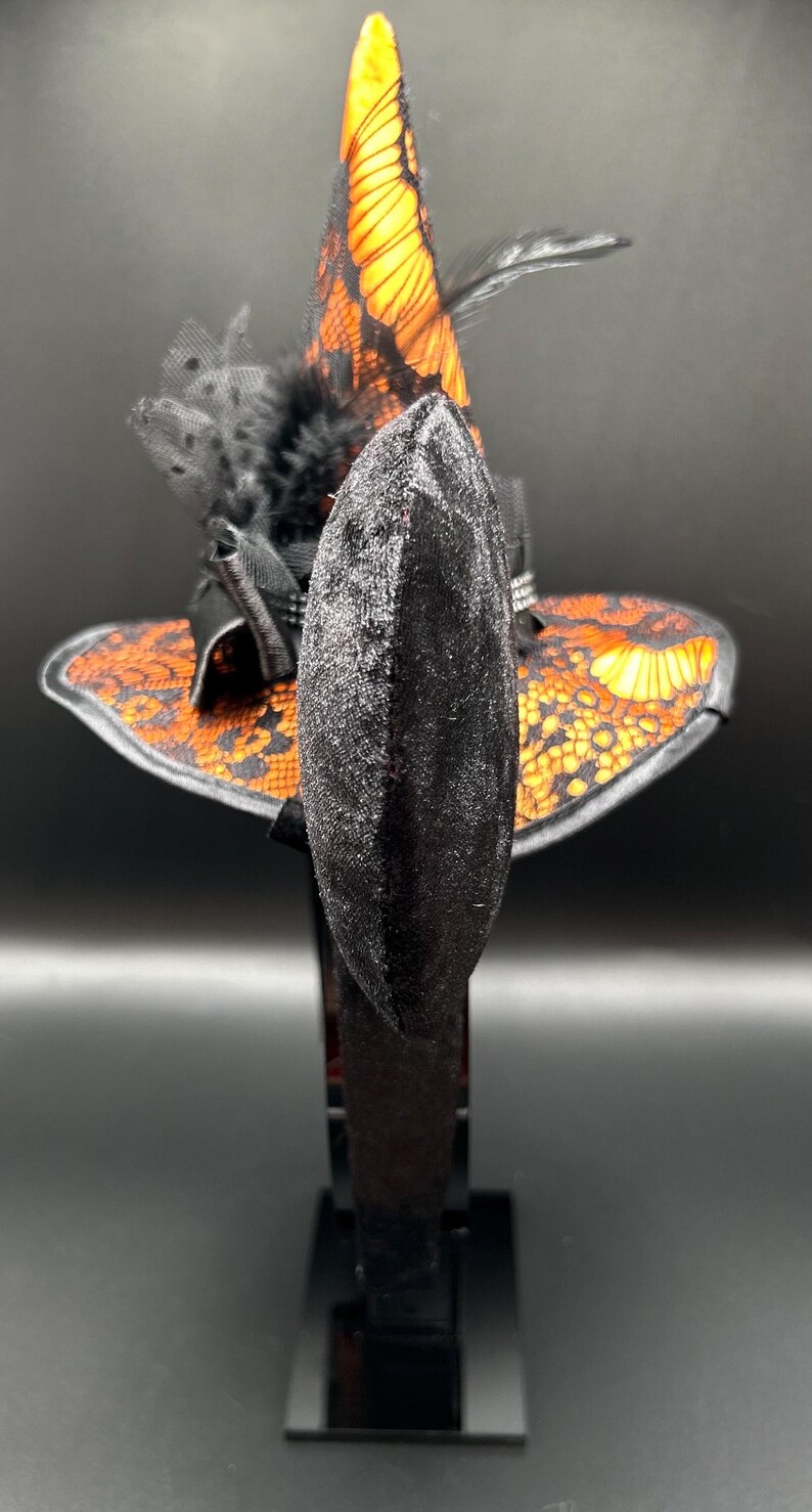 May include: A black velvet headband with a small witch hat design. The hat features orange and black lace detailing, a black feather, and a pointed top. The accessory is likely for Halloween or costume events.