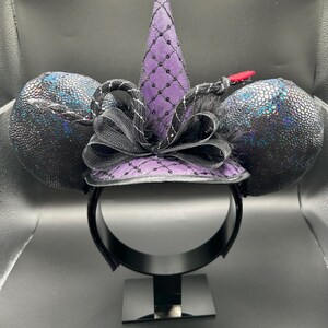 May include: Black and purple mouse ears headband with a witch hat design. The ears have a textured, iridescent finish. The hat is purple with a black net overlay and a black bow. A red accent is on the side.