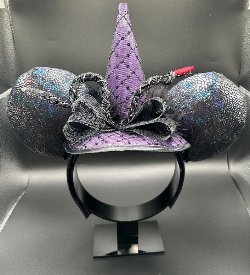 May include: Black and purple mouse ears headband with a witch hat design. The ears have a textured, iridescent finish. The hat is purple with a black net overlay and a black bow. A red accent is on the side.