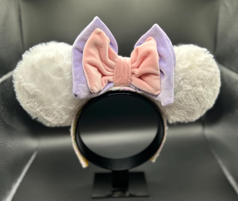 May include: White plush mouse ear headband with a lavender bow and pink center. The ears are fluffy and round, and the bow is made of soft velvet material. The headband is black.
