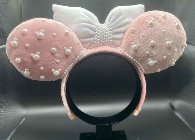 May include: Pink velvet mouse ears headband with a white bow and pearl embellishments.