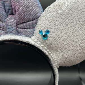 May include: White sequined mouse ears with a shimmering bow in shades of silver, blue, and pink. The ears feature a small, blue jewel design. The headband is black with a white rope trim.