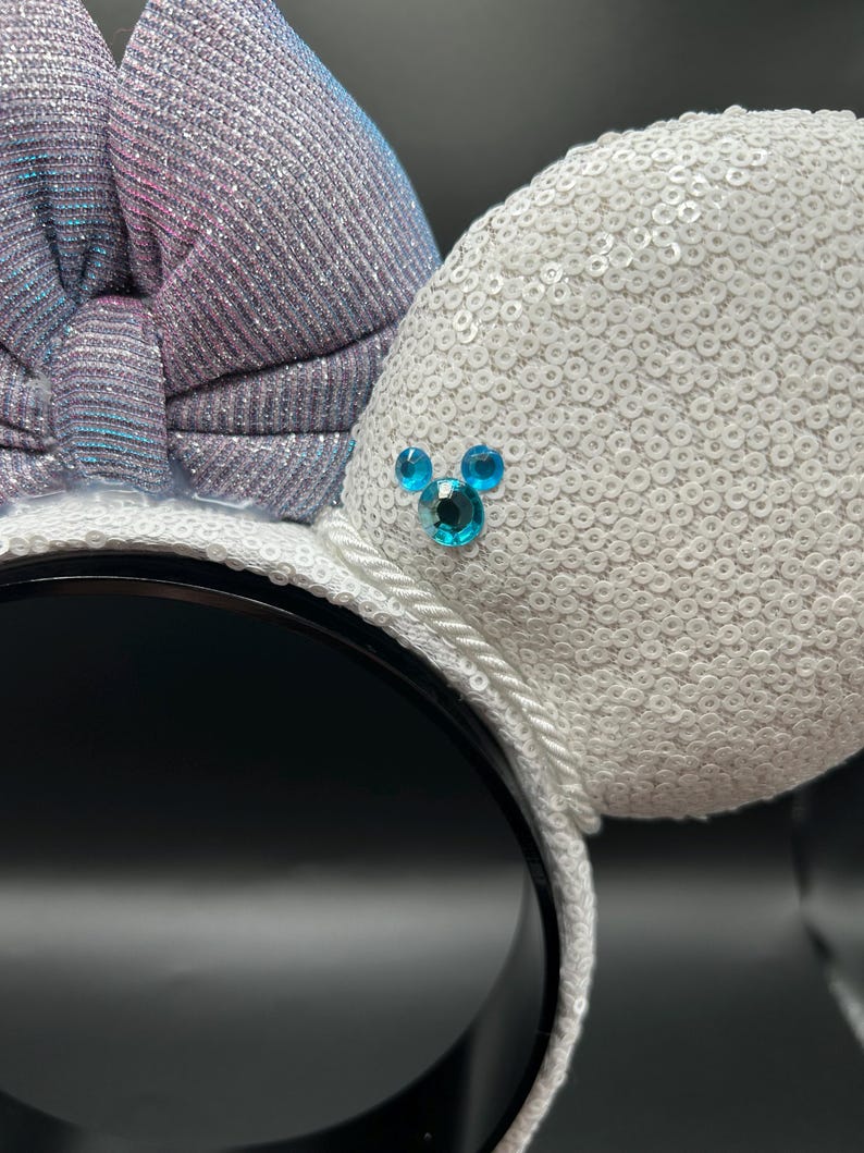 May include: White sequined mouse ears with a shimmering bow in shades of silver, blue, and pink. The ears feature a small, blue jewel design. The headband is black with a white rope trim.