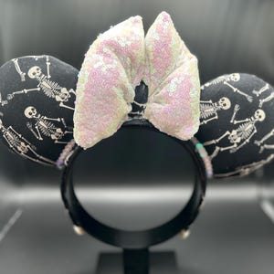 May include: Black mouse ears with a white skeleton print and a sequined bow. The ears are round and padded, with a black headband. The bow is a light pink color. The design is perfect for Halloween or a spooky theme.