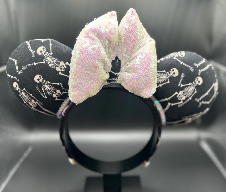 May include: Black mouse ears with a white skeleton print and a sequined bow. The ears are round and padded, with a black headband. The bow is a light pink color. The design is perfect for Halloween or a spooky theme.