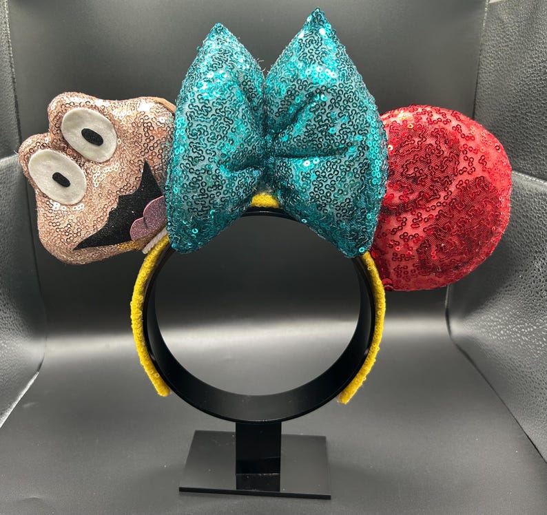 May include: A whimsical headband featuring a teal sequined bow, a red sequined ear, and a cartoon character ear. The headband is yellow and black, with a black base. The cartoon character ear is brown with white eyes and a black mouth.