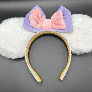 May include: A whimsical headband featuring fluffy white ears, a lavender and pink bow, and a yellow band adorned with sequins. The headband is designed for a fun and playful look, perfect for themed events or everyday wear.
