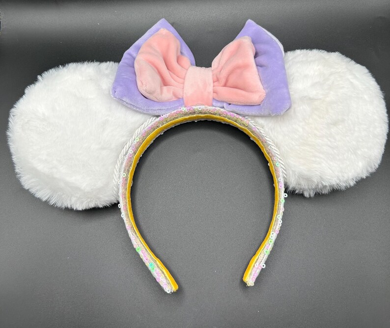 May include: A whimsical headband featuring fluffy white ears, a lavender and pink bow, and a yellow band adorned with sequins. The headband is designed for a fun and playful look, perfect for themed events or everyday wear.