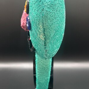 May include: A teal sequined headband with a rounded top, decorated with colourful accents. The headband features a row of sequins and is embellished with buttons and a pink, red, and yellow floral design. The accessory is displayed on a black stand.