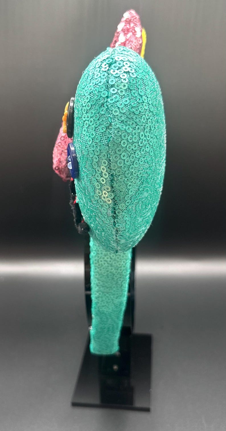 May include: A teal sequined headband with a rounded top, decorated with colourful accents. The headband features a row of sequins and is embellished with buttons and a pink, red, and yellow floral design. The accessory is displayed on a black stand.