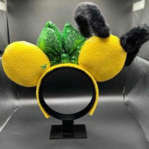 May include: A yellow headband with lemon-shaped accents and black fuzzy antennae. A green sequined bow sits between the lemon shapes. The headband is displayed on a black stand against a neutral background.