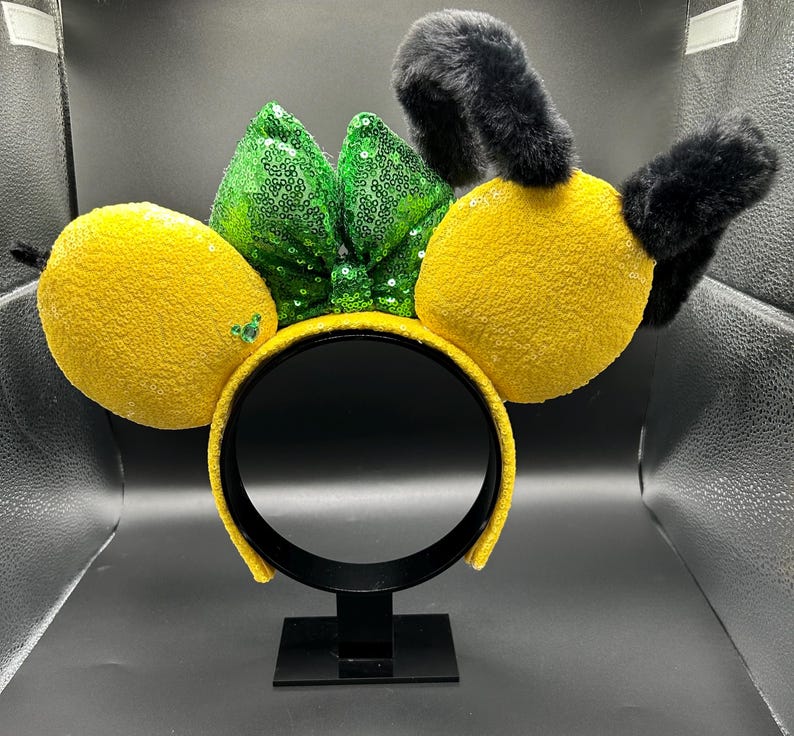 May include: A yellow headband with lemon-shaped accents and black fuzzy antennae. A green sequined bow sits between the lemon shapes. The headband is displayed on a black stand against a neutral background.