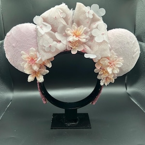 Op de afbeelding: Een haarband met twee ronde, lichtroze pluizige oren en een grote, centrale, glinsterende lichtroze strik. Het ontwerp is versierd met meerdere delicate, lichtroze kunstbloemen en kleine witte bloemblaadjes verspreid over de strik en de oren.
