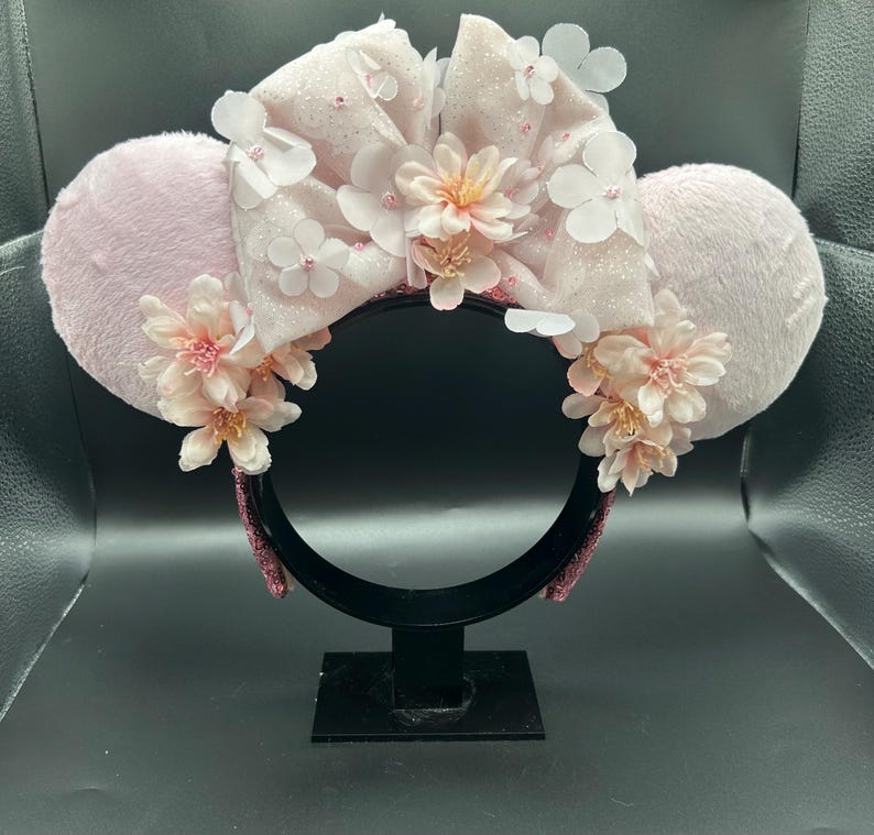 Op de afbeelding: Een haarband met twee ronde, lichtroze pluizige oren en een grote, centrale, glinsterende lichtroze strik. Het ontwerp is versierd met meerdere delicate, lichtroze kunstbloemen en kleine witte bloemblaadjes verspreid over de strik en de oren.