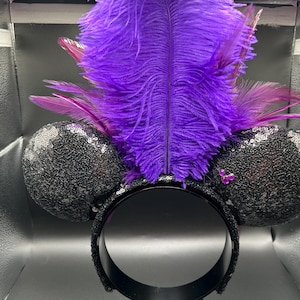 May include: A headband featuring two round black ears covered in shimmering sequins. A large, vibrant purple ostrich feather plume stands upright between the ears, accented by smaller purple feathers. A small cluster of three pink rhinestones is visible.