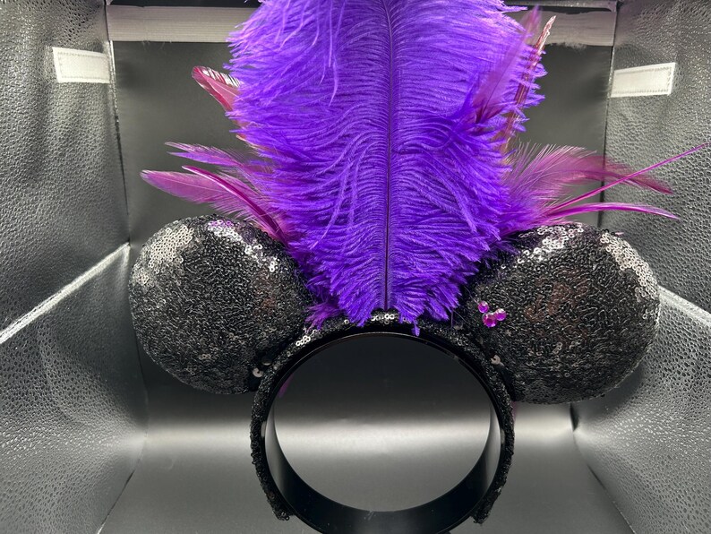 May include: A headband featuring two round black ears covered in shimmering sequins. A large, vibrant purple ostrich feather plume stands upright between the ears, accented by smaller purple feathers. A small cluster of three pink rhinestones is visible.