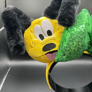 May include: A yellow headband featuring a Pluto character design. The headband has black, fluffy ears, white eyes, and a black nose. A green sequined bow is attached to the side, adding a touch of sparkle to this playful accessory.