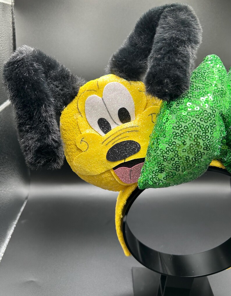 May include: A yellow headband featuring a Pluto character design. The headband has black, fluffy ears, white eyes, and a black nose. A green sequined bow is attached to the side, adding a touch of sparkle to this playful accessory.