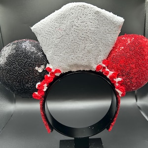 May include: A headband featuring two circular ears covered in black and red sequins, with a central white sequined crown-shaped piece. Small red and white fabric roses are arranged along the base of the ears and crown, and the headband is black.