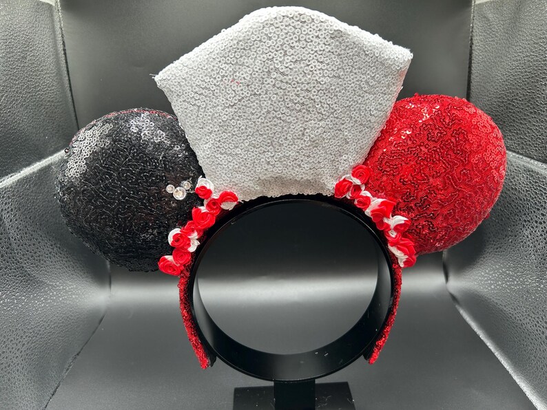 May include: A headband featuring two circular ears covered in black and red sequins, with a central white sequined crown-shaped piece. Small red and white fabric roses are arranged along the base of the ears and crown, and the headband is black.