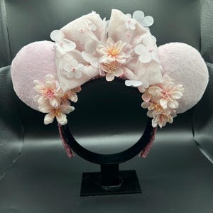 Op de afbeelding: Een haarband met twee ronde, lichtroze pluizige oren en een grote, glinsterende lichtroze strik in het midden. Het ontwerp is versierd met trossen zachtroze kunstbloemen en kleine witte bloemblaadjes, wat zorgt voor een gelaagd, bloemig uiterlijk.