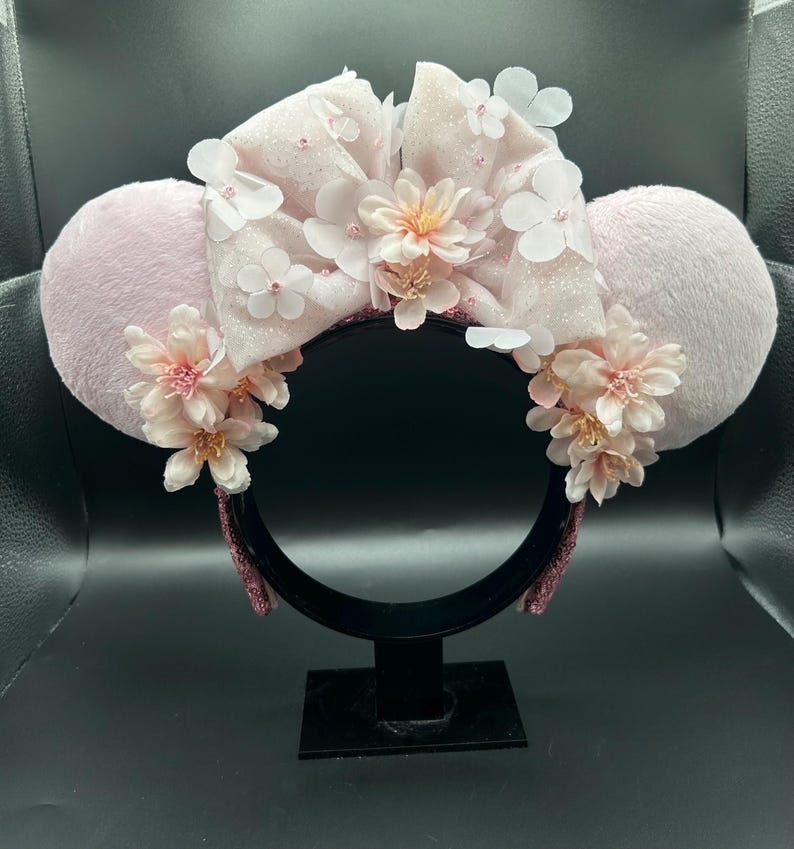 Op de afbeelding: Een haarband met twee ronde, lichtroze pluizige oren en een grote, glinsterende lichtroze strik in het midden. Het ontwerp is versierd met trossen zachtroze kunstbloemen en kleine witte bloemblaadjes, wat zorgt voor een gelaagd, bloemig uiterlijk.