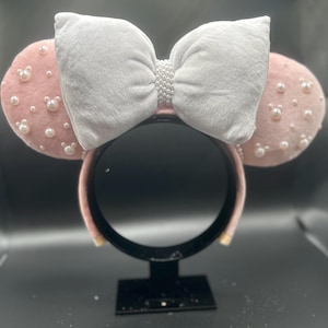 May include: Pink Minnie Mouse ears with a white velvet bow and pearl embellishments.