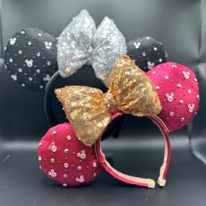 May include: Several decorative headbands with mouse ears and large sequined bows. One headband has black ears with rhinestones, another has silver sequins, and a third has gold sequins. A fourth headband has pink ears with rhinestones and small Mickey Mouse silhouettes.