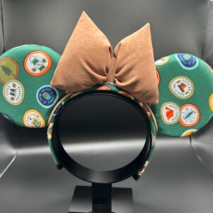 May include: Green mouse ears with a brown bow. The ears are covered in a pattern of colourful patches with camping-themed designs.