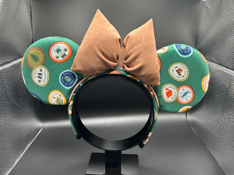 May include: Green mouse ears with a brown bow. The ears are covered in a pattern of colourful patches with camping-themed designs.