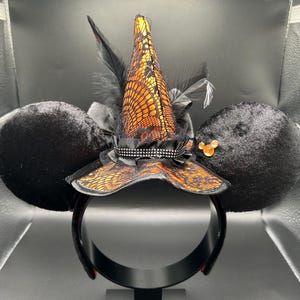 Puede incluir: Una diadema con dos grandes orejas redondas de terciopelo negro y un sombrero de bruja central. El sombrero es naranja con una capa de encaje negro, acentuado por plumas negras, una cinta negra y una peque&ntilde;a gema naranja en la oreja derecha.