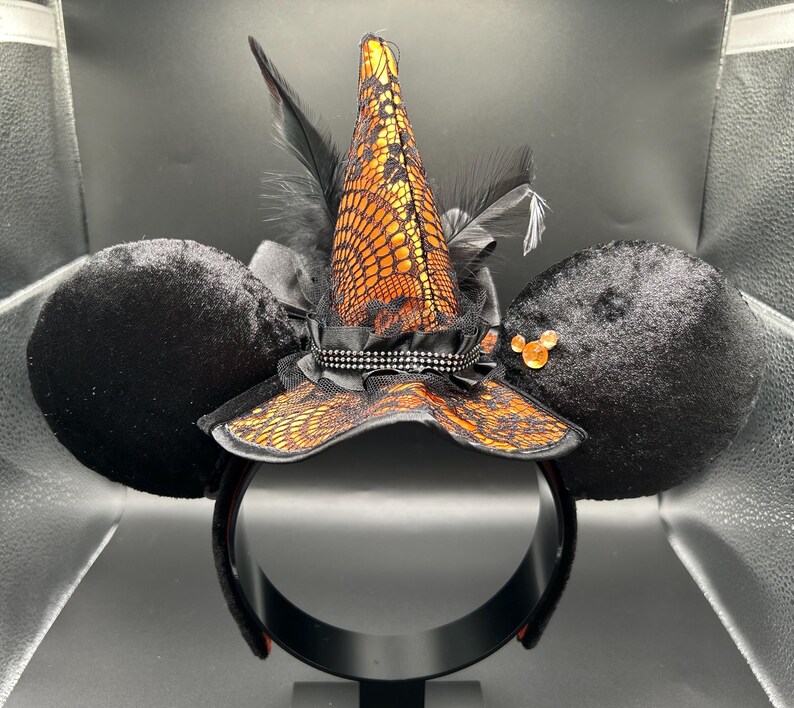 Puede incluir: Una diadema con dos grandes orejas redondas de terciopelo negro y un sombrero de bruja central. El sombrero es naranja con una capa de encaje negro, acentuado por plumas negras, una cinta negra y una peque&ntilde;a gema naranja en la oreja derecha.