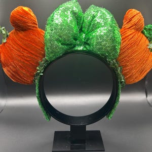 May include: A festive headband featuring orange, textured ears and a large, green sequined bow. The headband is adorned with small green leaves, creating a whimsical and playful accessory. The black headband provides a striking contrast to the bright colors.