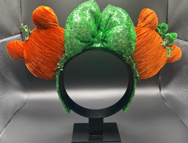 May include: A festive headband featuring orange, textured ears and a large, green sequined bow. The headband is adorned with small green leaves, creating a whimsical and playful accessory. The black headband provides a striking contrast to the bright colors.
