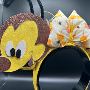 May include: Yellow and brown bee-themed Minnie Mouse ears with a honeycomb patterned bow. The ears are made of felt and have a 3D bee design.