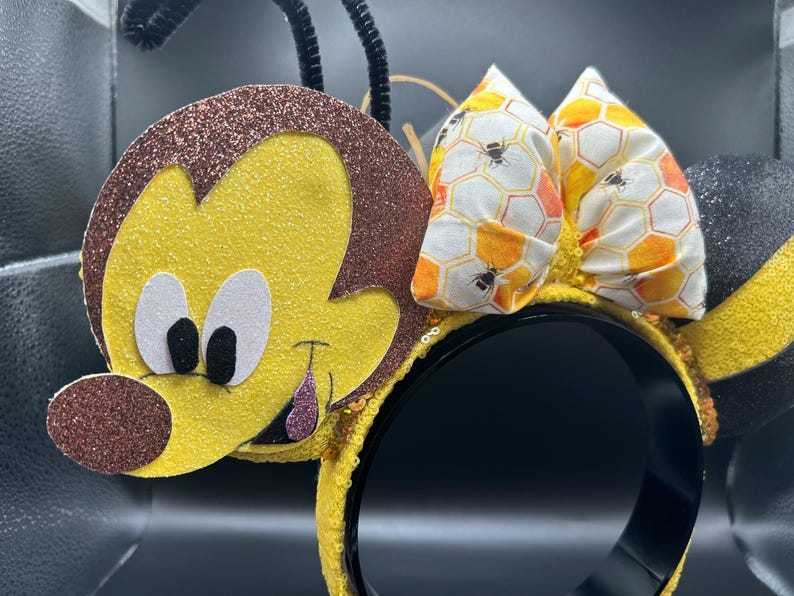 May include: Yellow and brown bee-themed Minnie Mouse ears with a honeycomb patterned bow. The ears are made of felt and have a 3D bee design.