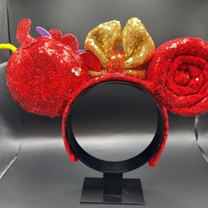 May include: Red sequined mouse ear headband with a gold sequined bow. The ears are round and covered in red sequins. A yellow pipe cleaner and purple accents add detail to the design.