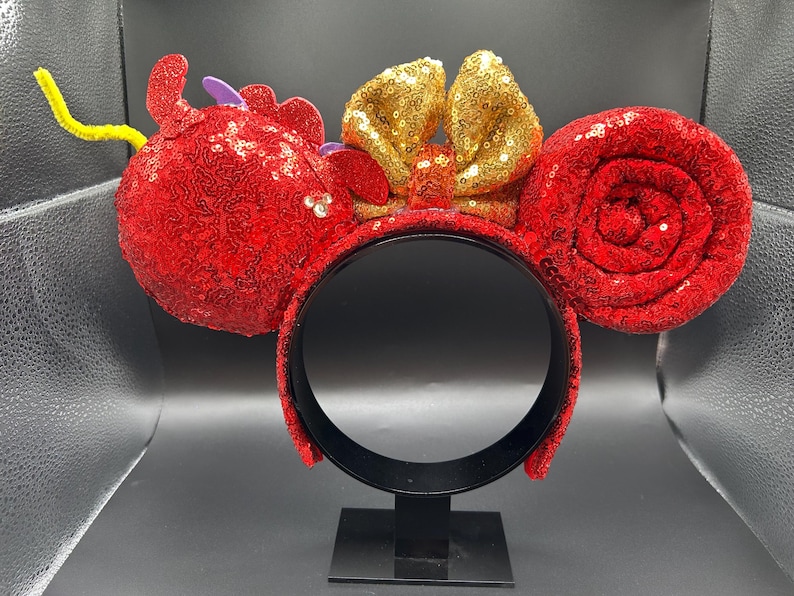 May include: Red sequined mouse ear headband with a gold sequined bow. The ears are round and covered in red sequins. A yellow pipe cleaner and purple accents add detail to the design.
