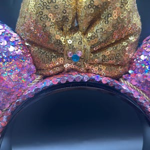 May include: A headband featuring two circular ears covered in iridescent pink sequins and a large central bow covered in gold sequins. The centre of the bow displays a small blue and pink icon shaped like a mouse head with rounded ears.
