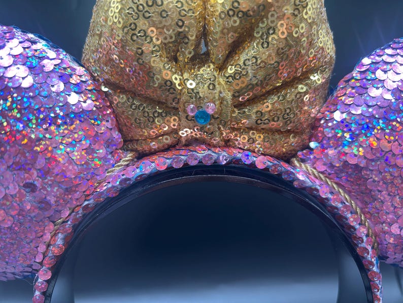 May include: A headband featuring two circular ears covered in iridescent pink sequins and a large central bow covered in gold sequins. The centre of the bow displays a small blue and pink icon shaped like a mouse head with rounded ears.
