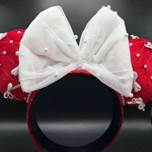 May include: Red and white Minnie Mouse ear headband. The ears are red with white bows and pearl accents. The headband is black with red sequins. The bow is made of white tulle and adorned with pearls.