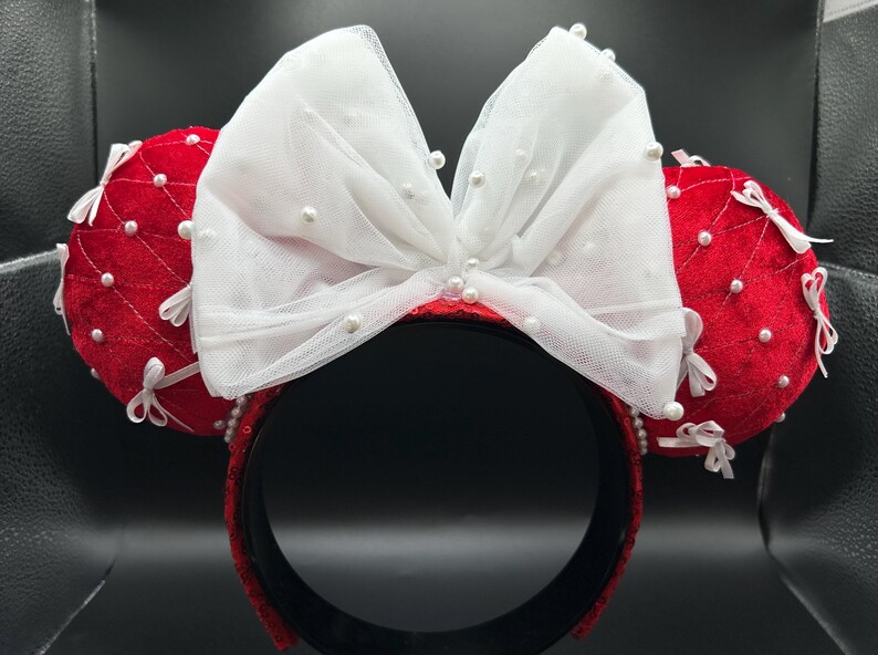 May include: Red and white Minnie Mouse ear headband. The ears are red with white bows and pearl accents. The headband is black with red sequins. The bow is made of white tulle and adorned with pearls.