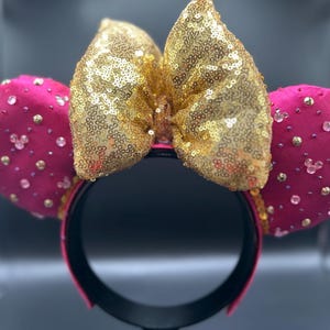 May include: A pair of Minnie Mouse ears with a gold sequined bow and hot pink ears. The ears are embellished with small gold and clear rhinestones. The headband is black with a pink trim.