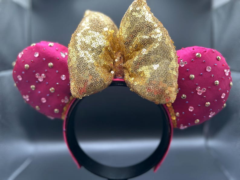 May include: A pair of Minnie Mouse ears with a gold sequined bow and hot pink ears. The ears are embellished with small gold and clear rhinestones. The headband is black with a pink trim.